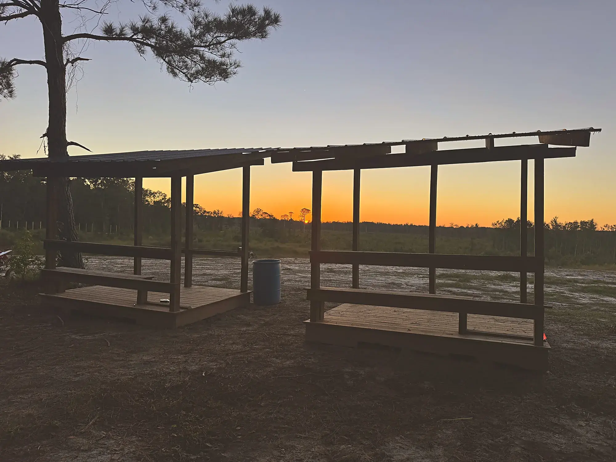 Two covered 5-stand shooting stations silhouetted against a Lowcountry sunset, with a tall pine and treeline behind