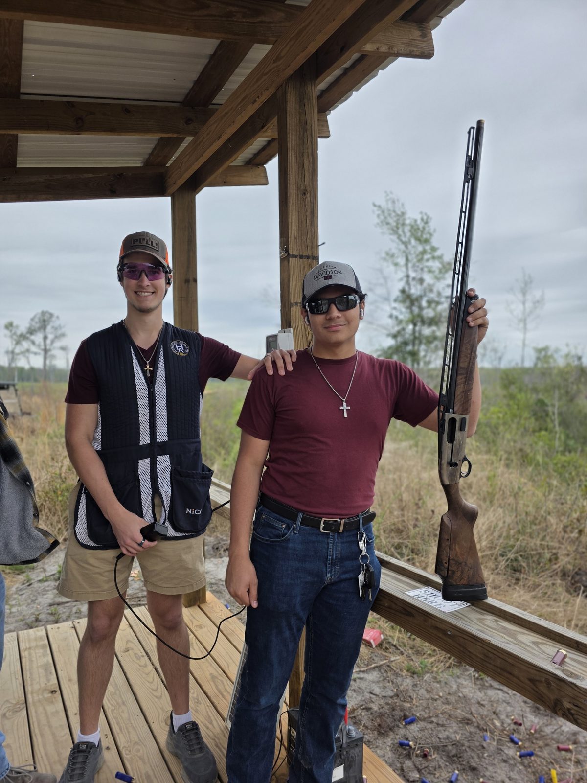 Two teen friends smiling under a shooting stand roof, one holding a shotgun pointed safely upward